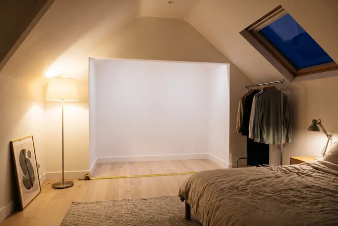 An empty alcove in a handsome, newly renovated attic bedroom in the evening, warmly lit, a tape measure stretched across the floor.