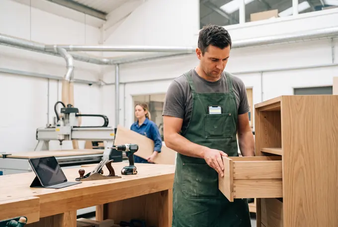 A joiner fits a drawer at his bench in a bright workshop, a phone lit in the chest pocket of his apron; a colleague carries a board past the CNC router.