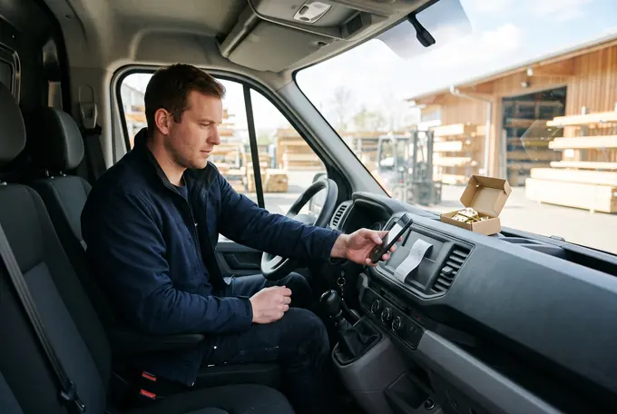 A joiner at the wheel of a new van photographs a till receipt with his phone; a box of brass hinges sits on the dashboard.